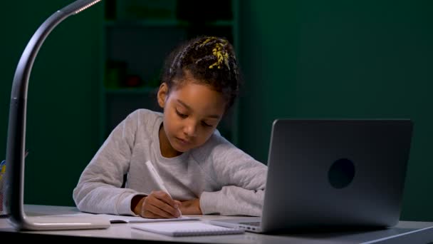 Una chica que usa una computadora portátil escucha un video tutorial y toma notas en su cuaderno. Un niño se sienta en una mesa en un cuarto oscuro a la luz de una lámpara de mesa. De cerca. Cámara lenta lista 59.97fps. — Vídeo de stock