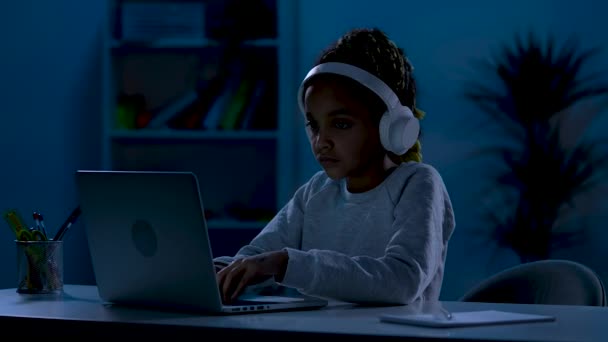 Una chica afroamericana seria con grandes auriculares blancos se sienta en una mesa en una habitación oscura, escribiendo en su computadora portátil. El concepto de aprendizaje a distancia. De cerca. Cámara lenta lista 59.97fps. — Vídeo de stock