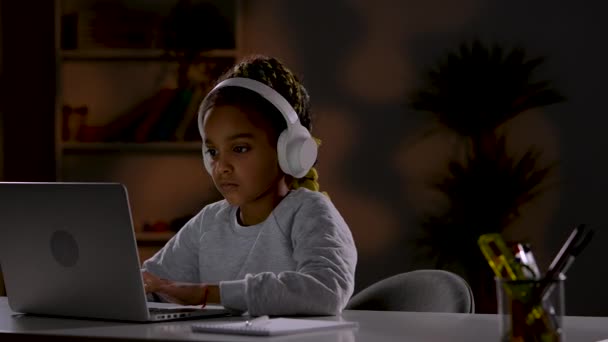 Una chica con auriculares inalámbricos blancos juega juegos en línea en su computadora portátil, sonríe y celebra una victoria. El niño se sienta en una mesa frente a un portátil en la habitación. De cerca. Cámara lenta lista 59.97fps. — Vídeos de Stock