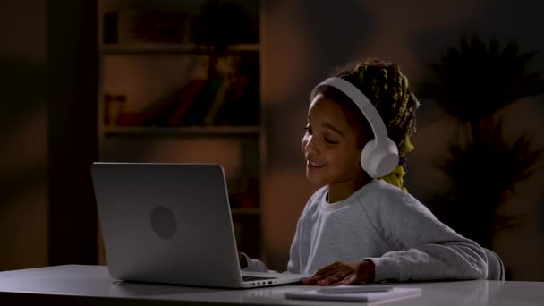 Beyaz kulaklıklı bir kız bilgisayarındaki bir video konuşmasında elini sallıyor, selamlıyor, canlı bir şekilde konuşuyor ve veda ediyor. Çocuk odadaki bir masada oturur. Kapatın. Yavaş çekim hazır 59.97fps. — Stok video