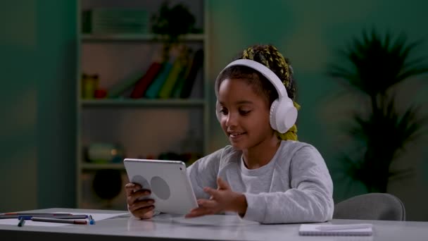 A girl in large white wireless headphones is talking on a video call on a tablet, waving her hand, greeting. The child sits at a table in the room. Close up. Slow motion ready 59.97fps. — Stock Video