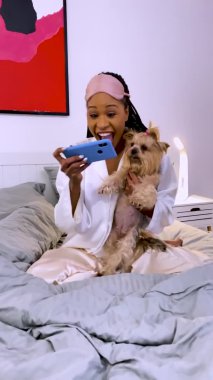 Bir kadın telefonuna bakar ve iyi haberlerle kutlar. Siyah bir kadın Yorkshire Terrier köpeğiyle yatak odasında pijamalarıyla oturuyor. Kapatın. Ağır çekim. Dikey video