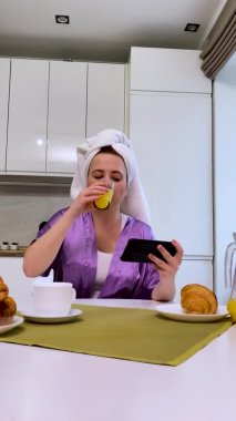 Genç ve güzel bir kadın mutfakta bir masaya oturur, meyve suyu içer, telefona bakar, iyi haberler alır, konuşur, aman Tanrım, zaferi kutlar. Ağır çekim. Dikey video.
