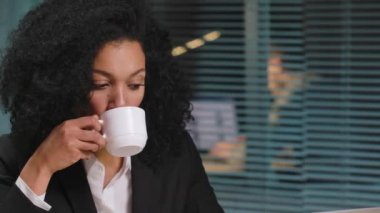 Afro-Amerikalı bir kadının portresi kahve içerken kafasını çeviriyor ve sorunu çözmeye çalışıyor. Ofisin içindeki masada oturan bir iş kadını. Kapatın. Yavaş çekim hazır 59.94fps.