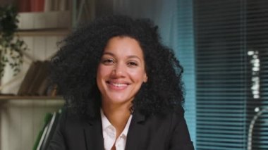İş elbisesi giymiş Afro-Amerikan kadın portresi kameraya bakıyor ve gülümsüyor. İş kadını iş yerinde poz veriyor, ofiste masada oturuyor. Kapatın. Yavaş çekim hazır 59.94fps.