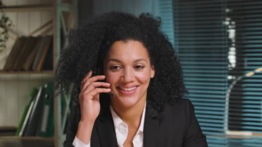 İş elbisesi giymiş Afro-Amerikan kadın portresi kulaklıkla, çağrı merkeziyle konuşuyor. İş kadını iş yerinde poz veriyor, ofiste masada oturuyor. Kapatın. Yavaş çekim hazır 59.94fps.