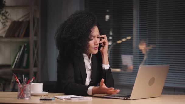 Портрет сердитої афроамериканської жінки на ноутбуці, яка розмовляє на смартфоні з обуренням. Зайнята жінка позувала на роботі, сидячи за столом у кабінеті. Повільний рух готовий до 59,94fps — стокове відео