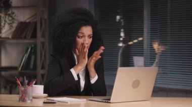 Afro-Amerikan kadın portresi dizüstü bilgisayardan heyecanlı bir şekilde konuşuyor, üzgün ve kafası karışık. İş kadını, iş yerinde poz veriyor, ofis içinde masada oturuyor. Yavaş çekim hazır 59.94fps.