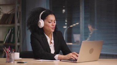 Afro-Amerikan kadın portresi büyük beyaz kulaklıklarla müzik dinliyor ve seviniyor. İş sırasında dinlenin. İş kadını iş yerinde poz veriyor, ofiste masada oturuyor. Yavaş çekim hazır 59.94fps.