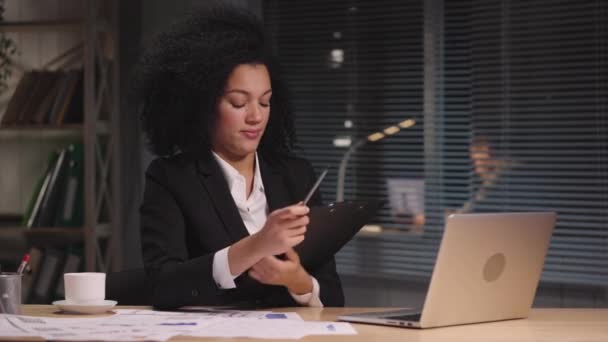 Retrato mujer afroamericana es charlas sobre videoconferencia en el ordenador portátil y muestra gráficos y diagramas. Empresaria posando en el lugar de trabajo, sentada a la mesa en la oficina. Cámara lenta lista 59.94fps. — Vídeo de stock