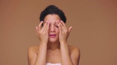 Genç Afro-Amerikan bir kadının güzel portresi parmaklarıyla yüzüne vuruyor ve nazik bir cilt masajı yapıyor. Siyah kadın, kahverengi stüdyo arka planında poz veriyor. Yavaş çekim hazır, 4K 59.94fps 'de..