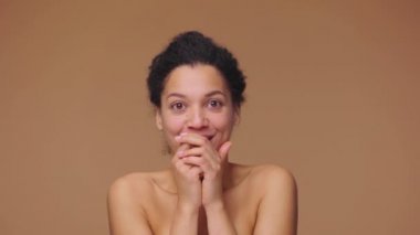 Genç Afro-Amerikalı bir kadın kameraya heyecanlı, keyifli ve etkileyici bir yüz ifadesiyle bakıyor. Kahverengi stüdyo arka planında siyah bir kadın. Yavaş çekim hazır, 4K 59.94fps 'de..