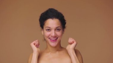 Afro-Amerikan genç bir kadının güzel portresi gülümseyen ve baş parmak hareketi gösteren. Kahverengi stüdyo arka planında poz veren siyah bir kadın model. Yavaş çekim hazır, 4K 59.94fps 'de..