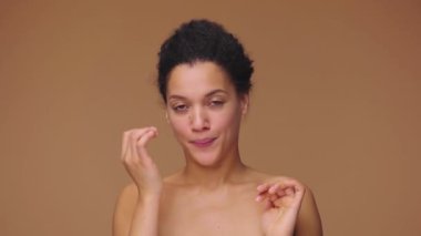Aynaya bakan, yüzüne dokunan ve saçlarını düzelten genç Afrikalı Amerikalı kadının güzel portresi. Kahverengi stüdyo arka planında poz veren siyah bir kadın model. Yavaş çekim hazır, 4K 59.94fps 'de..
