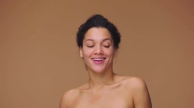 Genç Afrikalı Amerikalı kadının güzel portresi kameraya doğru dönüyor ve beyaz dişlerini göstererek gülümsüyor. Kahverengi stüdyo arka planında poz veren siyah bir kadın model. Yavaş çekim hazır, 4K 59.94fps 'de..