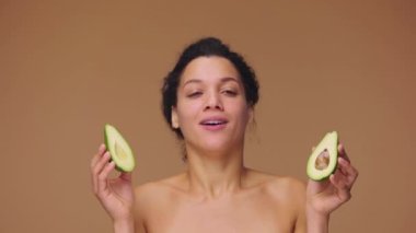Genç Afro-Amerikan bir kadının güzel portresi. Eğleniyor, gülümsüyor, avokado yarımlarını elinde tutuyor. Kahverengi stüdyo arka planında siyah bir kadın. Yavaş çekim hazır, 4K 59.94fps 'de..