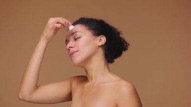 Yüzüne cilt serumu süren, parmaklarıyla nazikçe ovuşturan genç Afrikalı Amerikalı bir kadın. Siyah kadın, kahverengi stüdyo arka planında poz veriyor. Yavaş çekim hazır, 4K 59.94fps 'de..
