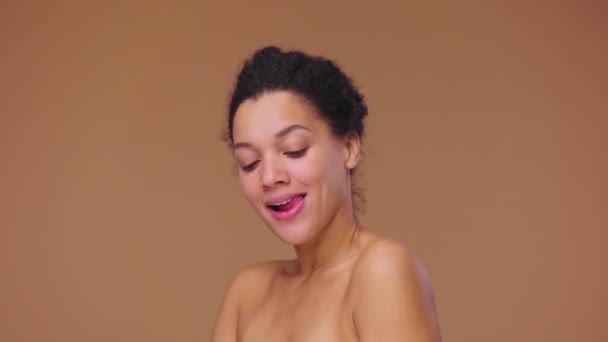 Schoonheidsportret van jonge Afro-Amerikaanse vrouw met een gezonde huid kijkend naar camera en koketterend glimlachend. Zwart model poserend op bruine studio achtergrond. Slow motion klaar, 4K bij 59,94fps — Stockvideo