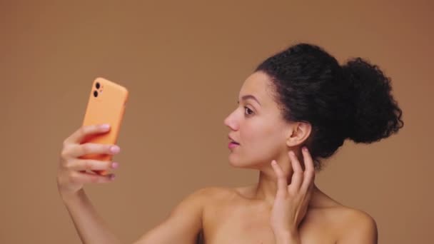 Piękny portret młodej Afroamerykanki robiącej selfie za pomocą smartfona. Czarna modelka pozująca na brązowym tle studia. Zwolnij, 4K przy 59,94fps.. — Wideo stockowe