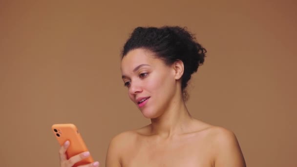 Retrato de belleza de una joven afroamericana que envía mensajes de texto en un teléfono inteligente y se regocija. Modelo femenino negro posando sobre fondo de estudio marrón. Cámara lenta lista, 4K a 59.94fps. — Vídeo de stock