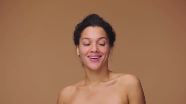 Skönhetsporträtt av ung afroamerikansk kvinna vänder sig mot kameran och ler, visar vita tänder. Svart kvinnlig modell poserar på brun studio bakgrund. Slow motion klar, 4K vid 59.94fps. — Stockvideo