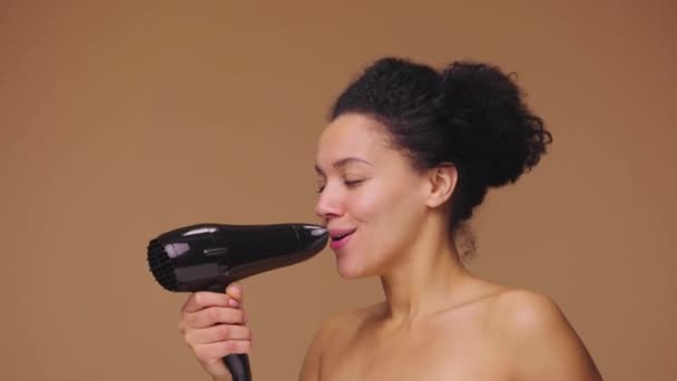 Beauty portrait of young African American woman dancing and singing merrily using hair dryer. Black female model posing on brown studio background. Slow motion ready, 4K at 59.94fps. — Stock Video