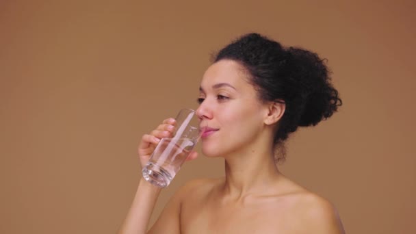 Schönheitsporträt einer jungen Afroamerikanerin, die Wasser aus Glas trinkt und eine Daumen-hoch-Geste zeigt. Schwarzes weibliches Modell posiert auf braunem Studiohintergrund. Zeitlupe bereit, 4K bei 59.94fps — Stockvideo