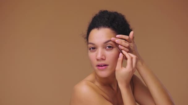 Retrato de belleza de una joven afroamericana mirándose en el espejo y arrancándose las cejas. Modelo femenino negro posando sobre fondo de estudio marrón. Cámara lenta lista, 4K a 59.94fps. — Vídeos de Stock