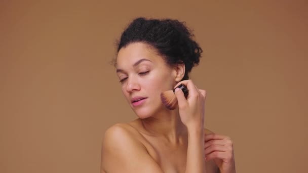 Schoonheidsportret van jonge Afro-Amerikaanse vrouw die poeder op haar gezicht aanbrengt met make-up borstel. Zwart vrouwelijk model poserend op bruine studio achtergrond. Slow motion klaar, 4K bij 59,94fps. — Stockvideo