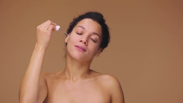 Beauté portrait jeune femme afro-américaine appliquant du sérum de soin sur le visage, frottant doucement avec les doigts. Femme noire posant sur fond de studio brun. Prêt au ralenti, 4K à 59.94fps. — Video