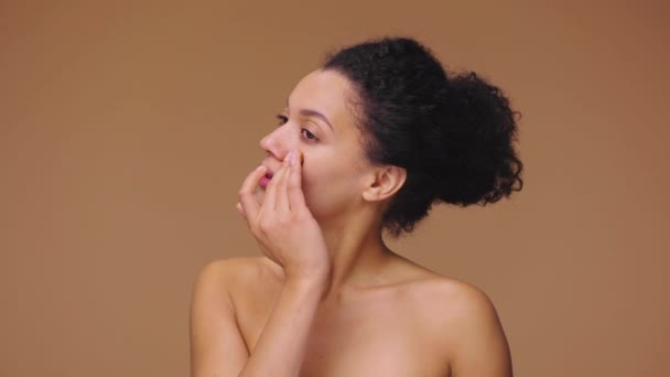 Portret de frumusețe tânără afro-americană care aplică scrub de îngrijire a pielii pe față, frecându-se ușor cu degetele. O femeie de culoare pozând pe fundalul studioului maro. Mișcare lentă gata, 4K la 59.94fps . — Videoclip de stoc