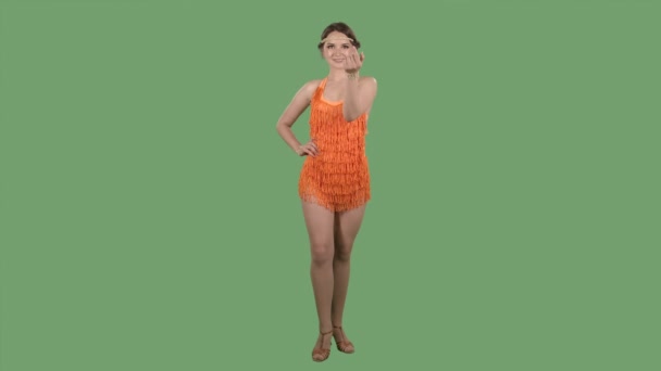 Krásná žena se dívá do kamery, koketně mává rukou a ukazuje gesto. Portrét brunetky v oranžových šatech s třásněmi na zelené obrazovce ve studiu. Zpomalený pohyb. — Stock video
