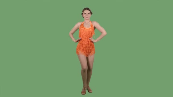 Portret van een mooie jonge vrouw die gracieus glimlachend en dansend naar de camera kijkt. Brandende brunette in een oranje franje jurk op een groen scherm in de studio. Langzame beweging. — Stockvideo