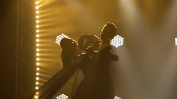 Professional couple of ballroom dancers waltzes on an iparket in dark studio against backdrop of bright lights. Man and woman dance partners are dancing at ball. Silhouettes. Close up. — Stock Video