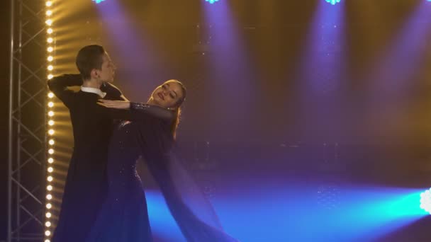 Элегантная пара танцовщиц танцующих вальс, прыгающих и держащихся за руки. Мужчина и женщина вальсируют в темной студии среди ярких синих прожекторов и дыма. Закрыть. — стоковое видео