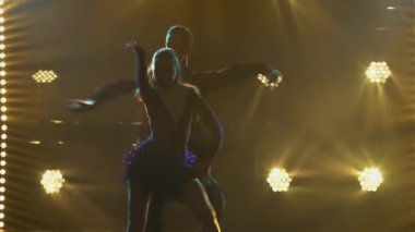 Rumba Drive dansı genç bir çift balo dansçısı tarafından gerçekleştiriliyor. Takım elbiseli bir adam ve kısa elbiseli bir kadın karanlık bir stüdyoda parlak ışıkların önünde neşeyle dans ediyorlar. Siluetler. Kapat..