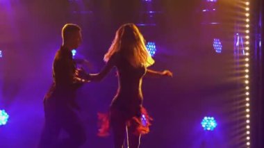 Tutkulu sarışın, rengarenk ışıkların önünde eşiyle rumba yapıyor. Karanlık bir stüdyoda Latin Amerika 'da dans eden çiftlerin siluetleri. Kapat..
