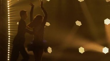 Uzun saçlı ince bir esmer partneriyle cha-cha-cha dansı yapıyor. Karanlık arka planda parlak ışıkları olan iki kişinin silueti. Kapat..