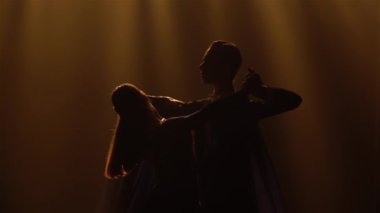 İki büyüleyici dansçı tarafından hafif vals hareketleri sergileniyor. Zarif takım elbiseli bir adam ve uzun elbiseli bir kadın sarı ışıkla karanlık stüdyoda vals yapıyor. Siluetler. Kapatın. Yavaş çekim.