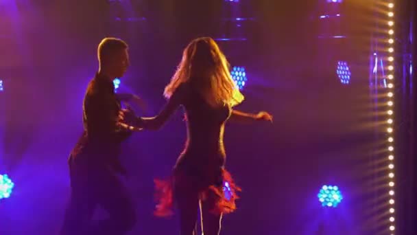 Страстная блондинка танцует румбу со своим партнером на фоне ярких огней. Силуэты пары танцоров, исполняющих элементы латиноамериканского бального танца в темной студии. Закрыть. — стоковое видео
