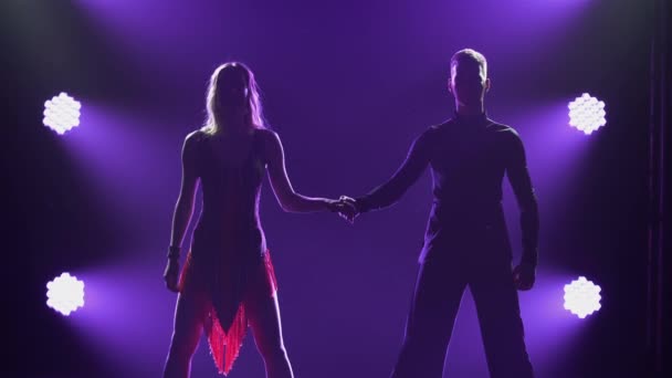Professionell föreställning av balsalsdansare i det latinamerikanska dansprogrammet. Silhuetter av sensuella par på en bakgrund av lila ljus i en mörk rökig studio. Närbild. — Stockvideo