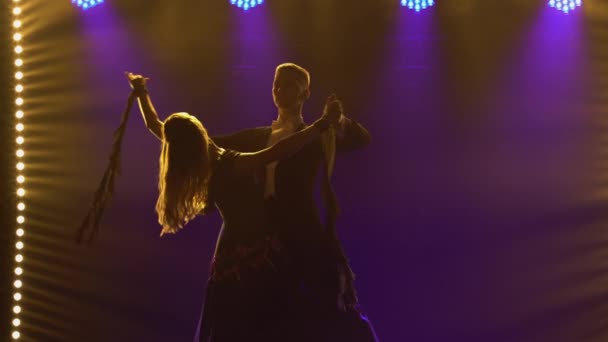 Elementy klasycznej choreografii walca w wykonaniu pary tancerzy towarzyskich. Partnerzy mężczyzna i kobieta walc trzymając się za ręce. Kręcony w ciemnym studio na czarnym tle z niebieskimi światłami. Zamknij się.. — Wideo stockowe