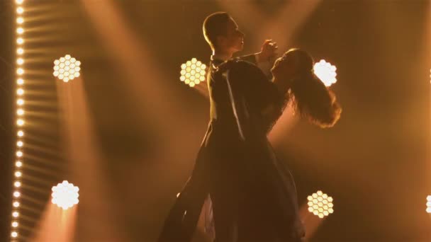 Silhouetted pair of ballroom dancers practicing dancing jumping holding hands. Classical dance choreography in dark studio against backdrop of multicolored spotlights. Close up. Slow motion. — Stock Video