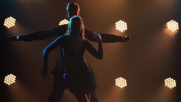 Rumba drive dance performed by pair of ballroom dancers. Man in suit and woman in short dress are dancing merrily in dark studio against background of bright lights. Silhouettes. Close up. Slow motion — Stock Video