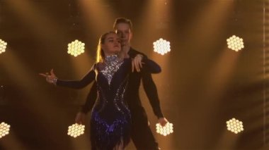 Klasik balo salonu Latin Amerika dansının unsurları profesyonel dansçılar tarafından sahneleniyor. Rumba, samba, cha-cha-cha. Erkekler ve kadınlar sarı ışıklarla karanlık stüdyoda yakın dans ediyorlar. Yavaş çekim.