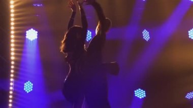 Uzun saçlı ince bir esmer partneriyle cha-cha-cha dansı yapıyor. Siyah arka planda parlak spot ışıklarıyla iki kişinin silueti. Kapatın. Yavaş çekim.