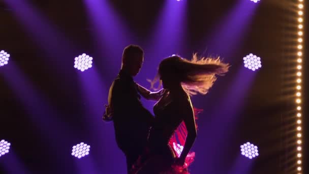 Actuación profesional de bailarines de salón en el programa de danza latinoamericana. Siluetas de pareja sensual sobre un fondo de luces moradas en un oscuro estudio ahumado. De cerca. Movimiento lento. — Vídeo de stock