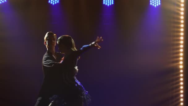 Jeune beau couple dansant des éléments de danse sensuelle cha-cha-cha, rumba, samba. Silhouettes de danseurs dans un studio sombre avec projecteurs bleus. Ferme là. Mouvement lent. — Video