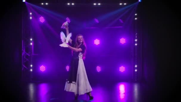 Young attractive woman performs with trained white doves in dark studio against the backdrop of blue lights. Birds sit in the hands of the trainer and flap their wings. Circus show with bird tricks. — Stock Video