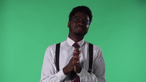 Egy fiatal afro-amerikai férfi portréja, amint mosolyog, valami érdekeset mond, és a beszélgetőpartnerre mutat. Fekete férfi nyakkendővel és fehér inges szemüveggel, zöld vásznon pózol a stúdióban. Közelről.. — Stock videók